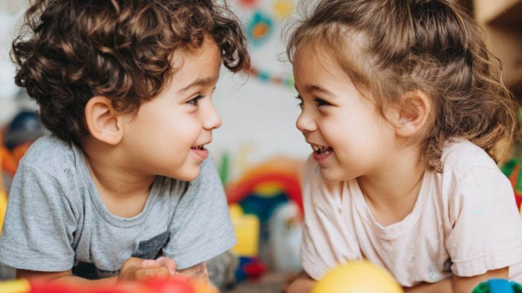 Niño y niña pequeños jugando juntos en la escuela infantil, sonriendo mientras se miran rodeados de juguetes de colores.