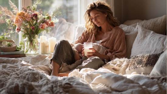 Mujer en recuperación posparto sentada en la cama con su bebé recién nacido, sosteniendo un vaso de bebida, rodeada de flores y luz suave que entra por la ventana.