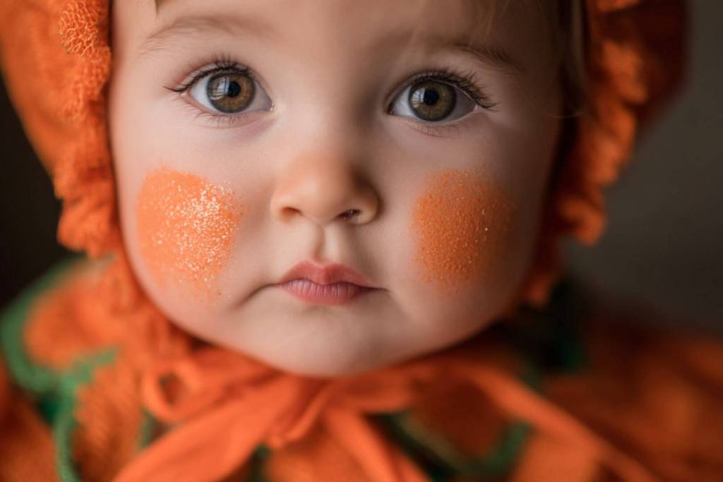 Primer plano de un bebé con disfraz de calabaza y maquillaje naranja en el rostro.