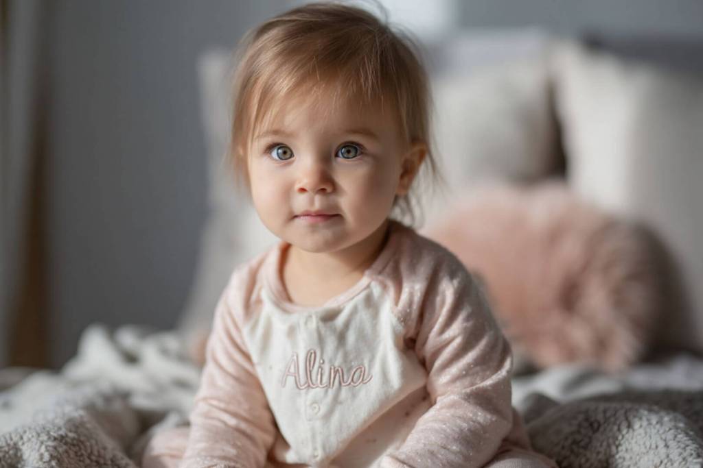 Bebé niña sentada sobre una cama con el nombre Alina bordado en el body