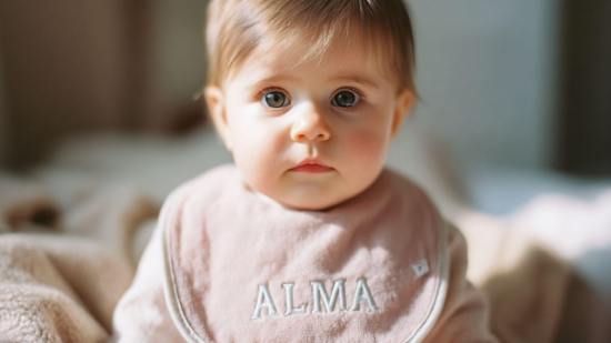 Bebé de ojos grandes con babero rosa donde se lee ALMA, en ambiente acogedor.