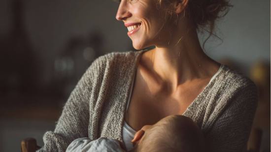 Mujer joven amamantando a su bebé en casa, con luz cálida y expresión sonriente, transmitiendo calma y ternura.