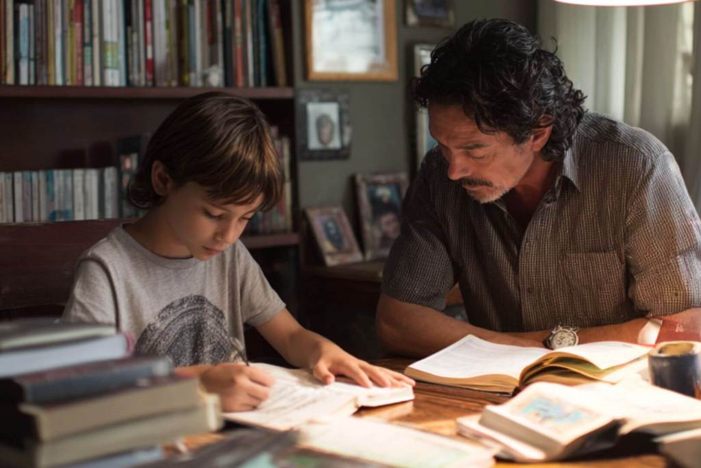 Niño haciendo deberes junto a su padre en casa, rodeados de libros.