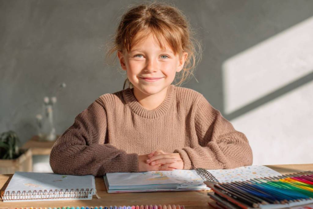 Niña sonriente sentada en la mesa organizando cuadernos y lápices de colores.