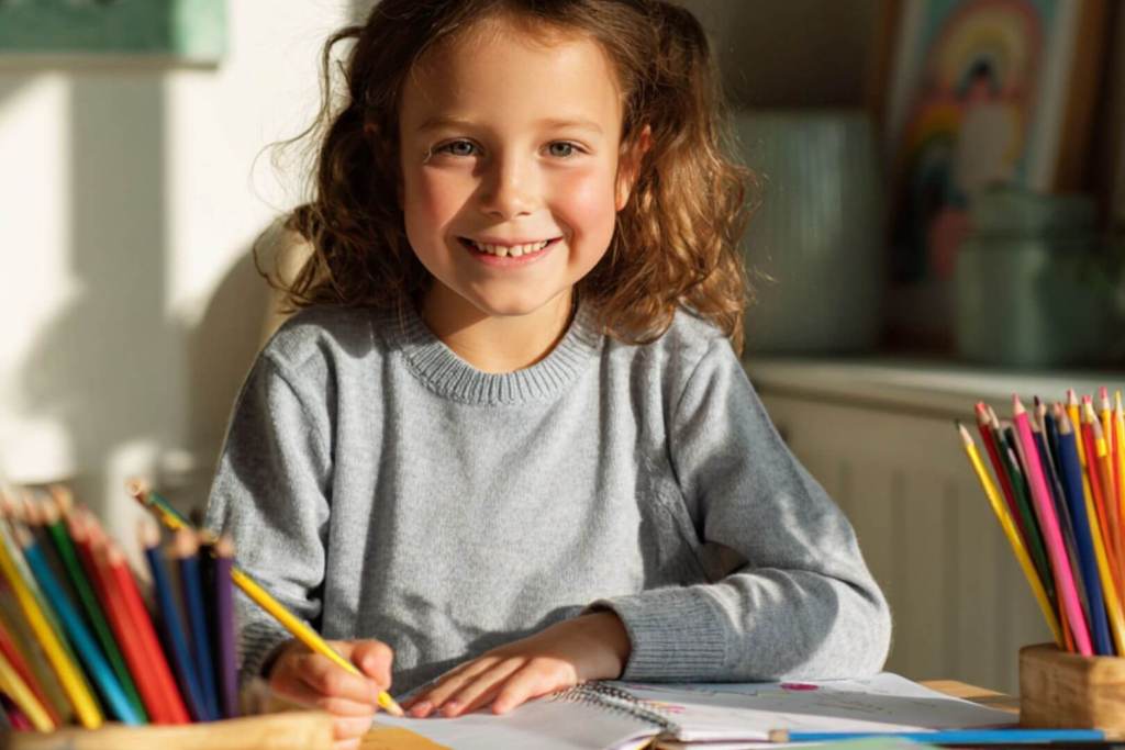 Niña con cuadernos y lápices ordenados sobre la mesa mientras sonríe a la cámara.