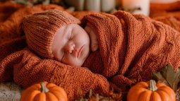 Recién nacido envuelto en manta naranja, rodeado de calabazas y velas en una escena cálida de Halloween.