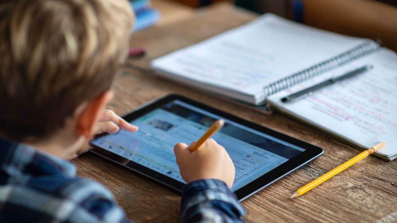 Niño en un pupitre escolar usando una tablet mientras toma apuntes en un cuaderno.