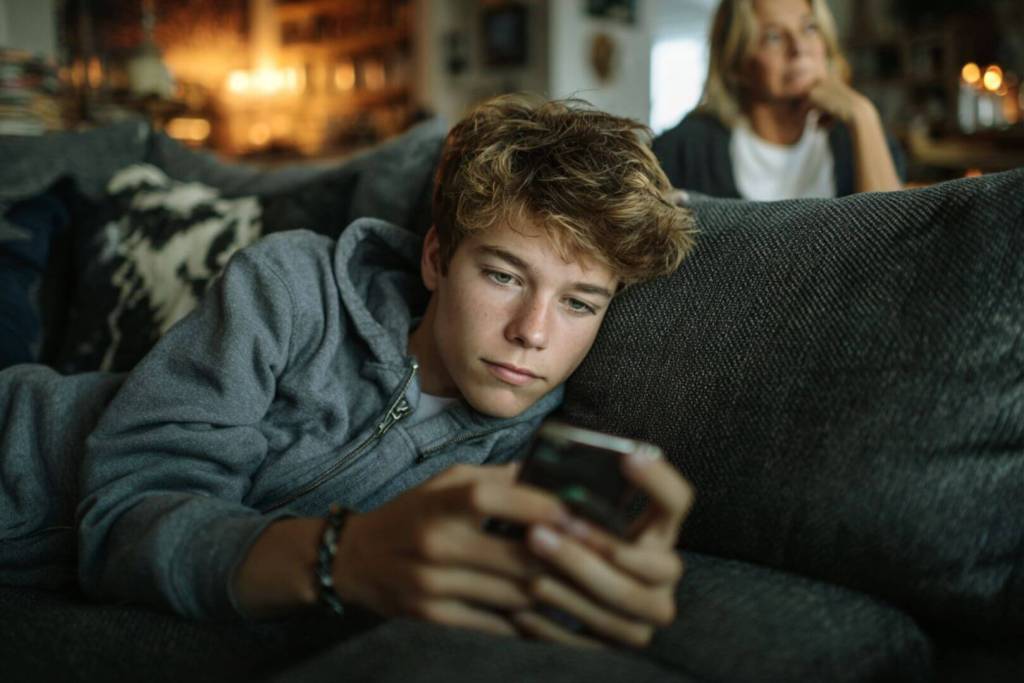 Un adolescente usa su smartphone mientras su madre lo observa en segundo plano, en un ambiente relajado del hogar.