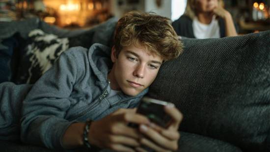 Un adolescente usa su smartphone mientras su madre lo observa en segundo plano, en un ambiente relajado del hogar.
