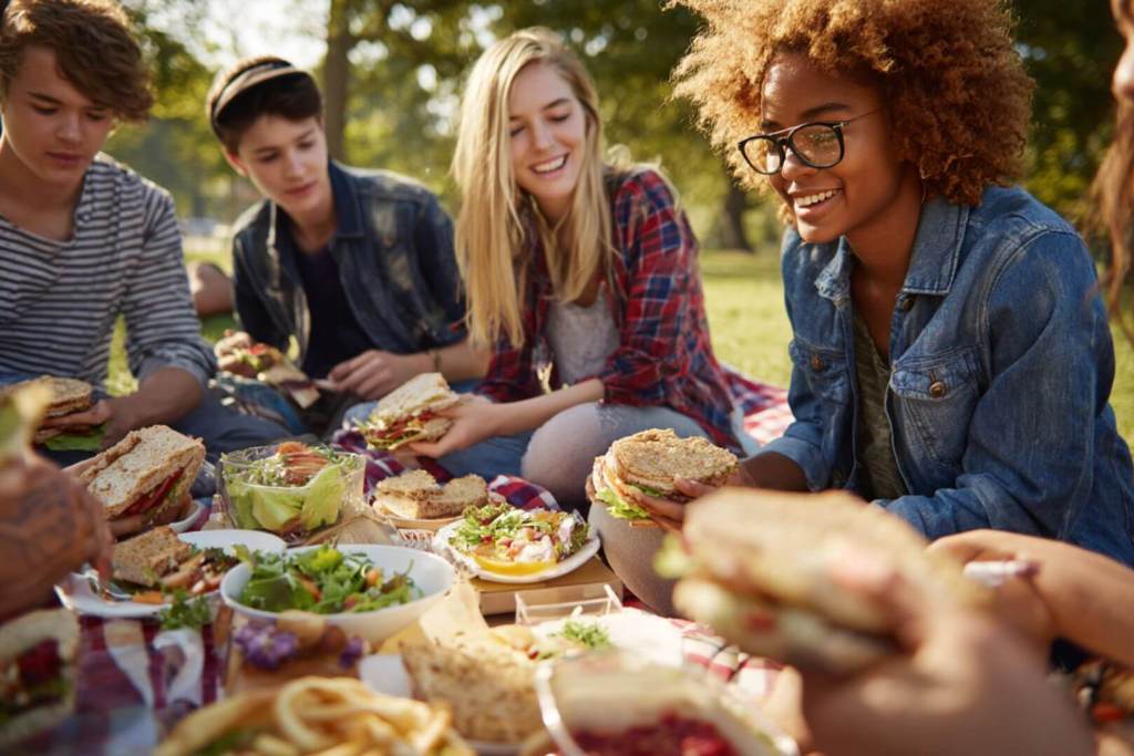 Adolescentes sonriendo mientras comparten una comida saludable con ensaladas, frutas y botellas de agua en un comedor escolar.