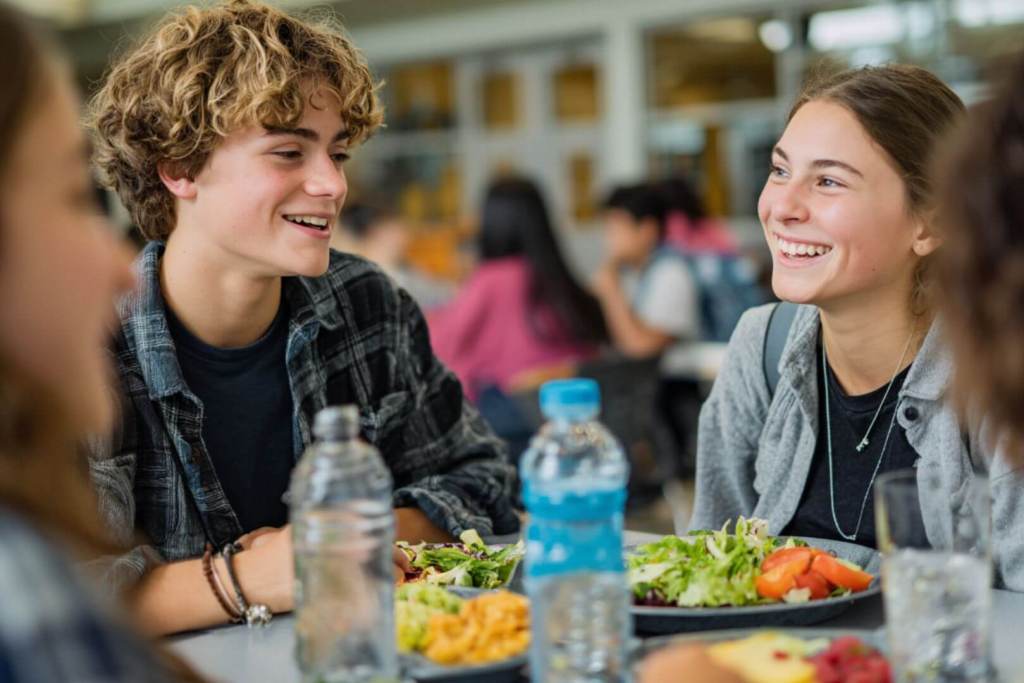 Dos adolescentes conversan alegremente mientras almuerzan ensaladas frescas y beben agua en un entorno escolar.