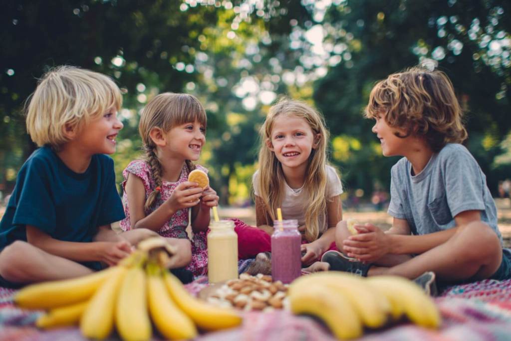 Cuatro niños disfrutando de batidos y frutas frescas en un picnic al aire libre.