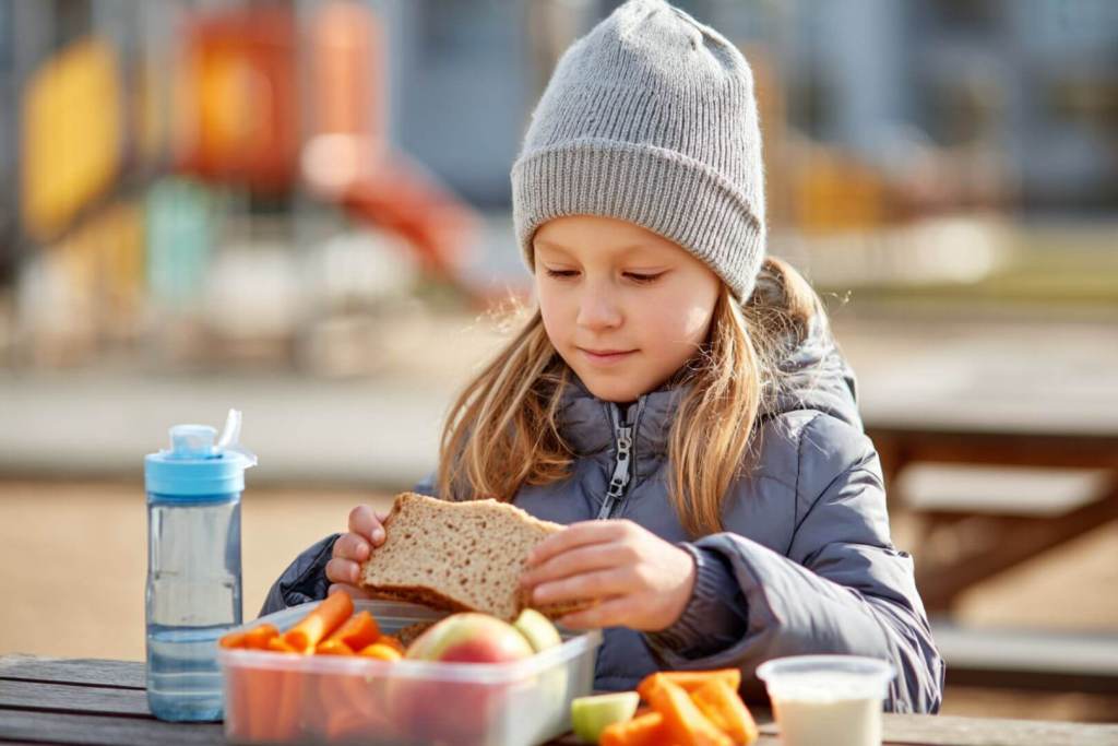 Niña con gorro de lana comiendo un bocadillo y verduras en un parque.