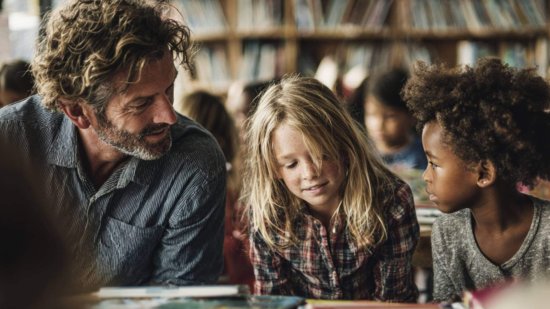 Maestro en aula moderna conversa con dos niños que leen libros en inglés, rodeados de materiales escolares y estanterías llenas de cuentos.