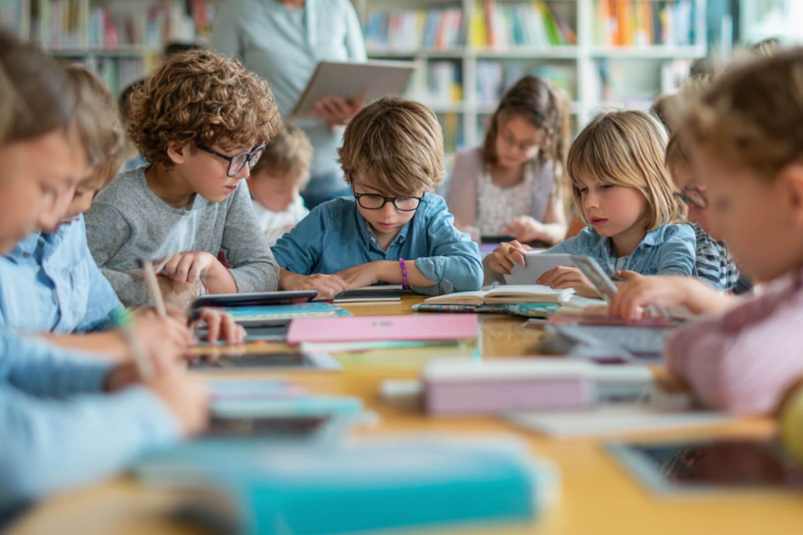 Niños de primaria estudiando juntos en clase, usando libros, cuadernos y tablets en una mesa compartida.