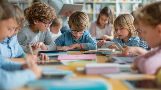 Niños de primaria estudiando juntos en clase, usando libros, cuadernos y tablets en una mesa compartida.