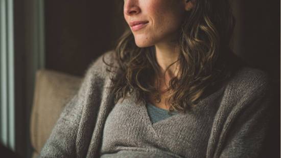 Mujer embarazada con suéter gris, sentada junto a una ventana, mirando pensativa hacia afuera, con luz natural suave iluminando su rostro.