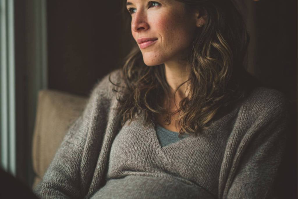 Mujer embarazada con suéter gris, sentada junto a una ventana, mirando pensativa hacia afuera, con luz natural suave iluminando su rostro.