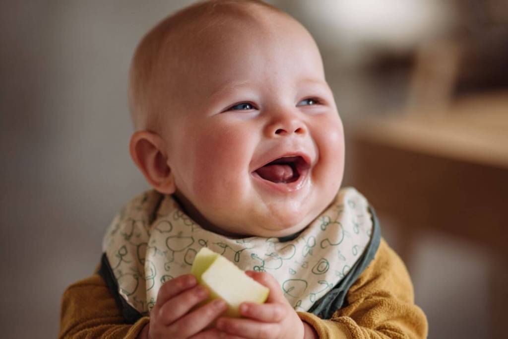Bebé sonriente con babero rosa sosteniendo una rodaja de manzana.