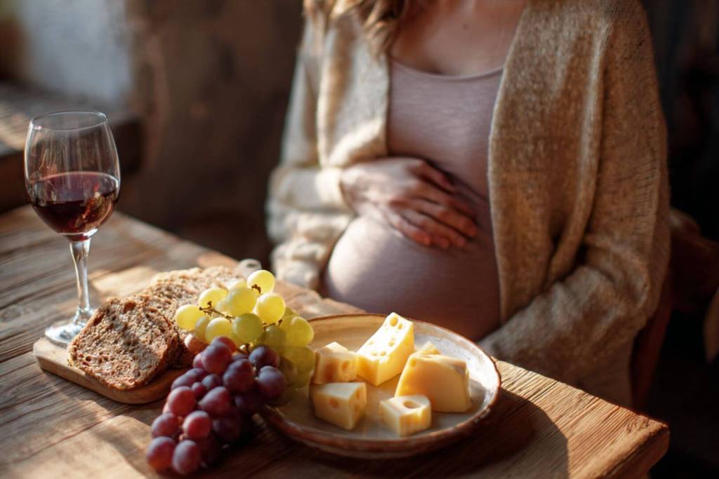 Mujer embarazada frente a un plato con quesos, uvas y pan integral junto a una copa de vino.