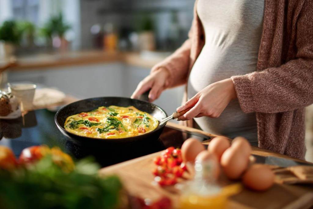 Mujer embarazada preparando una tortilla con vegetales frescos en la cocina.