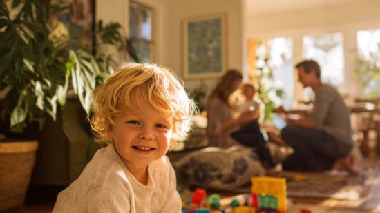 Niño pequeño jugando en casa con bloques de colores mientras sus padres cuidan al bebé en un ambiente familiar y luminoso.