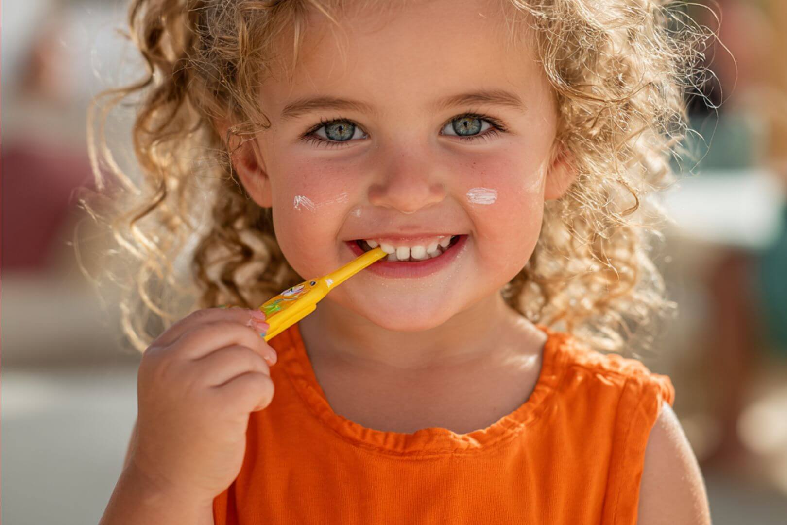 Niña pequeña con crema solar en las mejillas y cepillo de dientes en la mano, sonriendo mientras cuida su higiene y salud diaria.