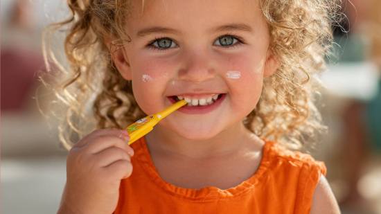 Niña pequeña con crema solar en las mejillas y cepillo de dientes en la mano, sonriendo mientras cuida su higiene y salud diaria.