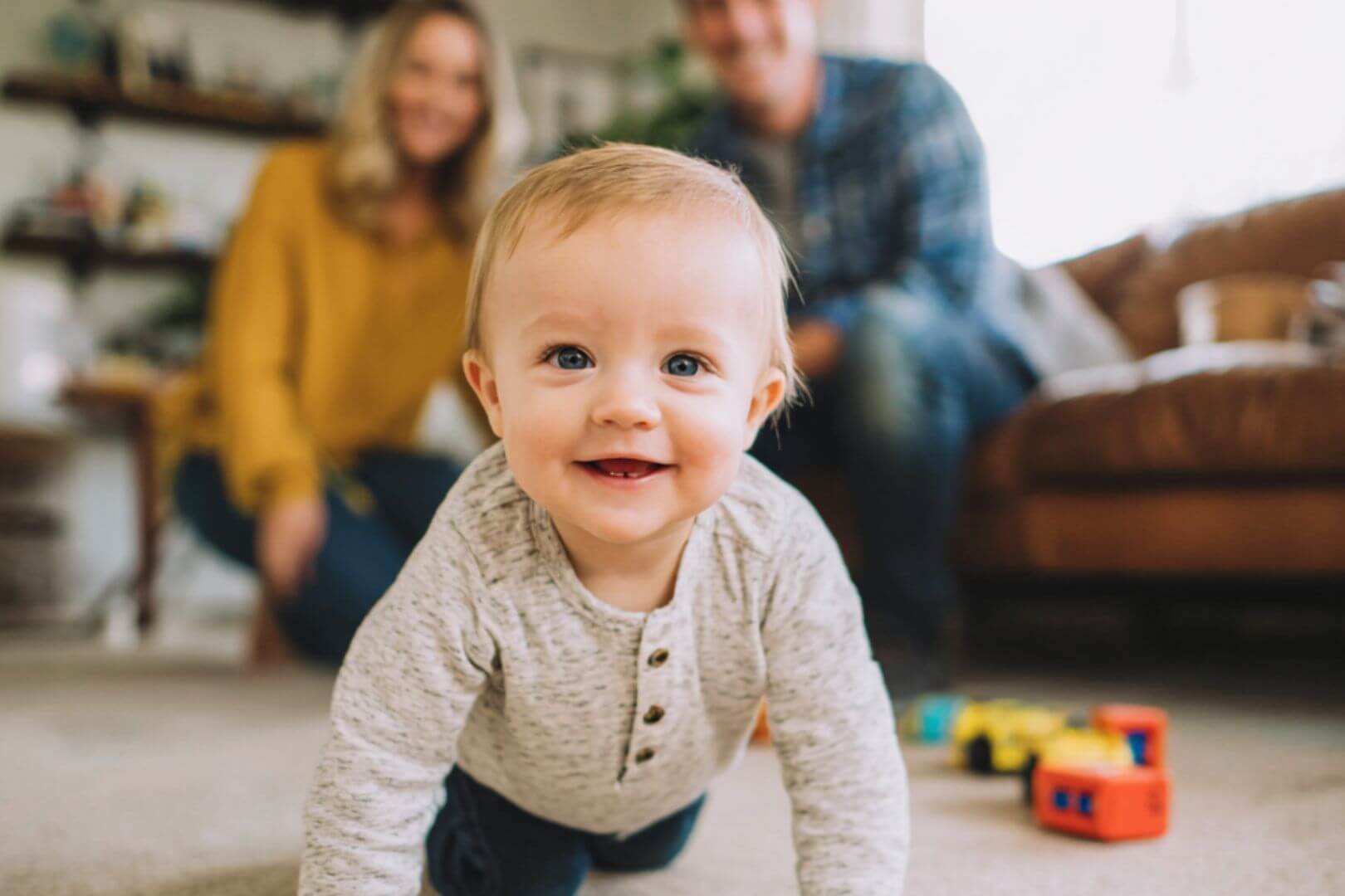 Bebé gateando y sonriendo, con sus padres al fondo en un ambiente familiar acogedor.