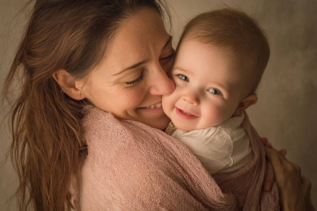 Madre abrazando y sonriendo a su bebé que mira a cámara.