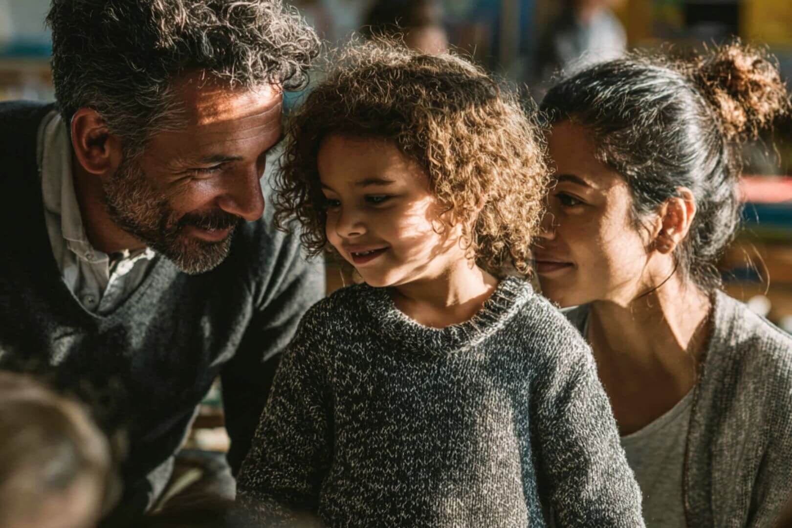 Madre y padre sonríen mientras apoyan a su hija pequeña en un espacio educativo, mostrando cercanía y afecto en su aprendizaje.