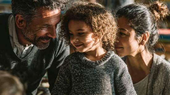 Madre y padre sonríen mientras apoyan a su hija pequeña en un espacio educativo, mostrando cercanía y afecto en su aprendizaje.
