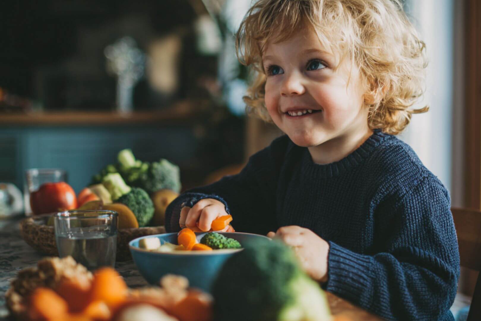 Niño pequeño sonriente sentado a la mesa mientras come verduras frescas como zanahorias y brócoli en un ambiente hogareño.