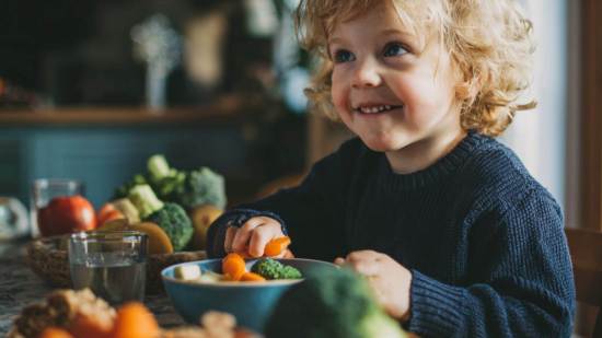 Niño pequeño sonriente sentado a la mesa mientras come verduras frescas como zanahorias y brócoli en un ambiente hogareño.