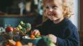 Niño pequeño sonriente sentado a la mesa mientras come verduras frescas como zanahorias y brócoli en un ambiente hogareño.