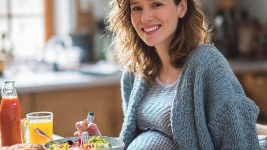 Mujer embarazada sonriendo mientras come una ensalada colorida y bebe jugo natural, sentada en la cocina con luz natural suave