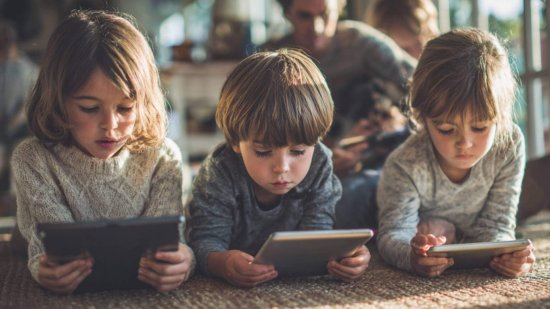 Tres niños sentados en el suelo mirando tablets con concentración, usando aplicaciones educativas en un entorno familiar luminoso.
