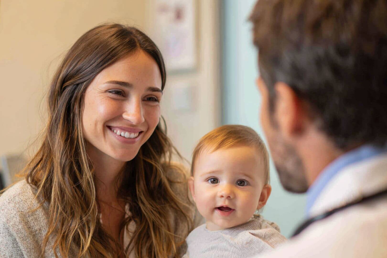 Madre sonriente sostiene a su bebé mientras habla con el pediatra en una consulta médica.