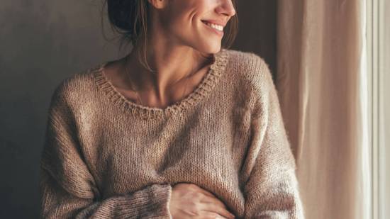 Mujer embarazada con suéter de punto mirando por la ventana y sonriendo, con una mano sobre su vientre en un gesto de ternura.