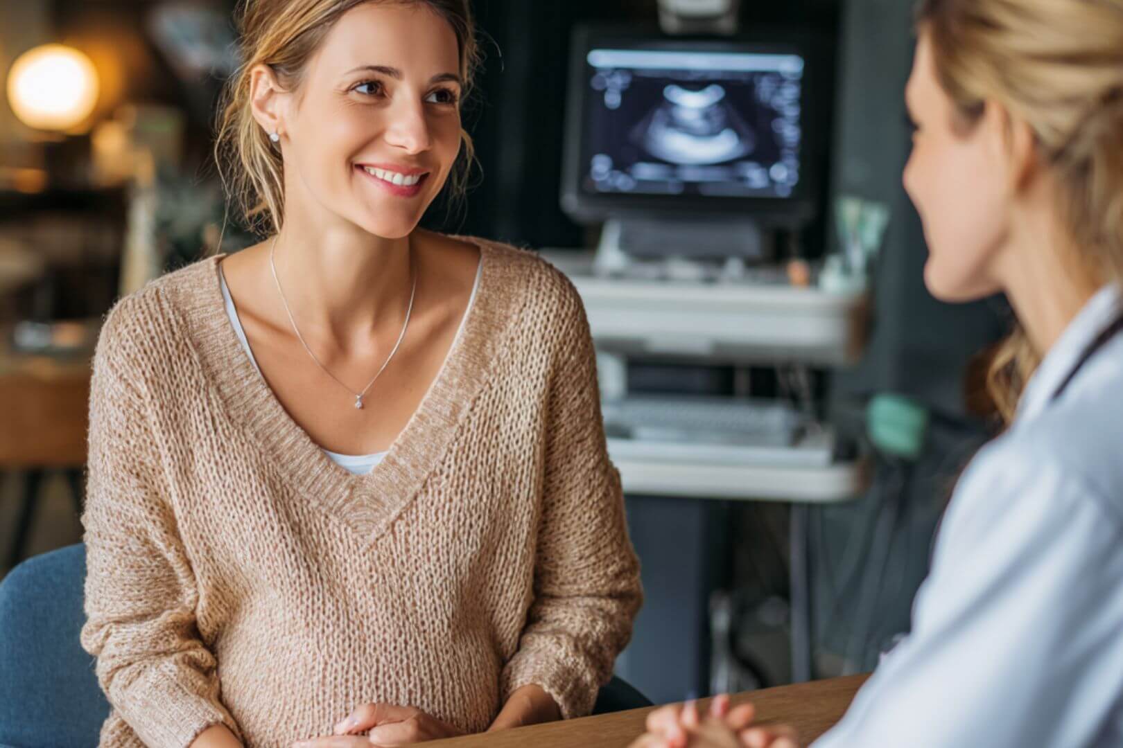 Mujer embarazada sonriente en consulta médica, con ecógrafo y pantalla mostrando imagen prenatal al fondo.