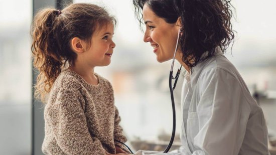 Niña en la consulta del pediatra mientras la doctora le escucha el corazón con un estetoscopio.