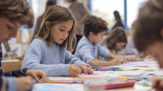 Grupo de alumnos trabajando en una actividad en clase, concentrados en sus mesas, mientras una maestra supervisa al fondo.