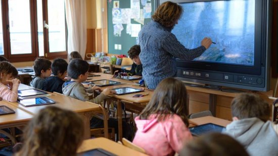 Aula de primaria en España con un profesor enseñando en pizarra digital y niños usando tablets en sus mesas.