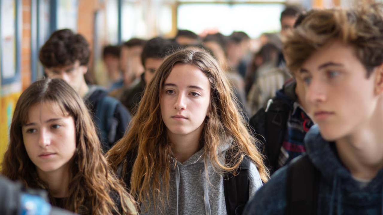Adolescentes españoles en un pasillo de instituto, con mochilas y expresiones serias.