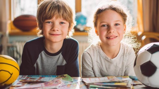 Dos niños sonríen en casa mientras eligen entre diferentes actividades extraescolares, rodeados de balones y folletos.