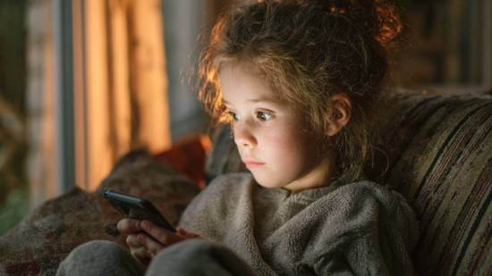 Niña pequeña sentada en el sofá mirando fijamente la pantalla de un teléfono móvil en casa.