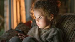 Niña pequeña sentada en el sofá mirando fijamente la pantalla de un teléfono móvil en casa.