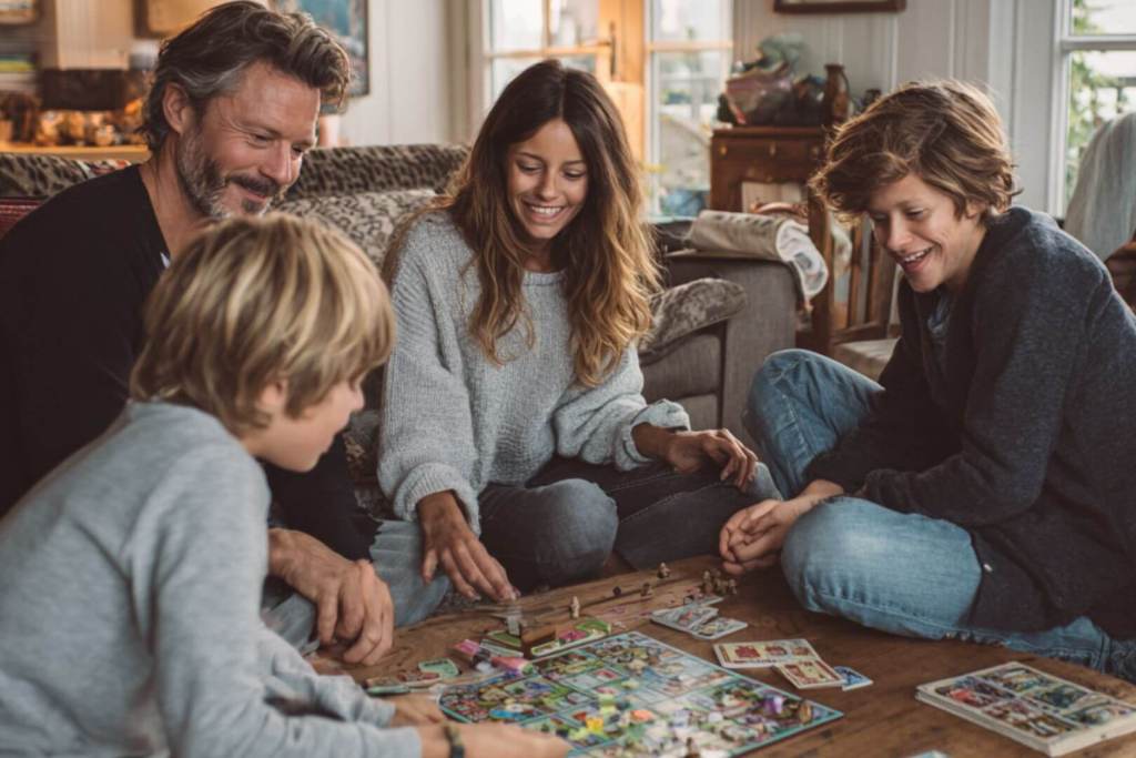 Padres y dos hijos adolescentes juegan a un juego de mesa en el salón, compartiendo tiempo de ocio en familia con risas y complicidad.