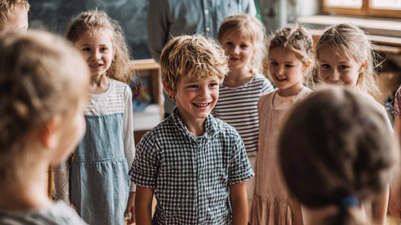 Grupo de niños de pie en el aula con su profesor al fondo, participando en una dinámica de movimiento frente a la pizarra.