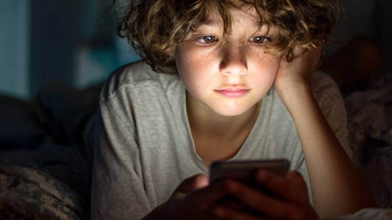 Niño tumbado en la cama mirando la pantalla iluminada de un teléfono móvil en la oscuridad.